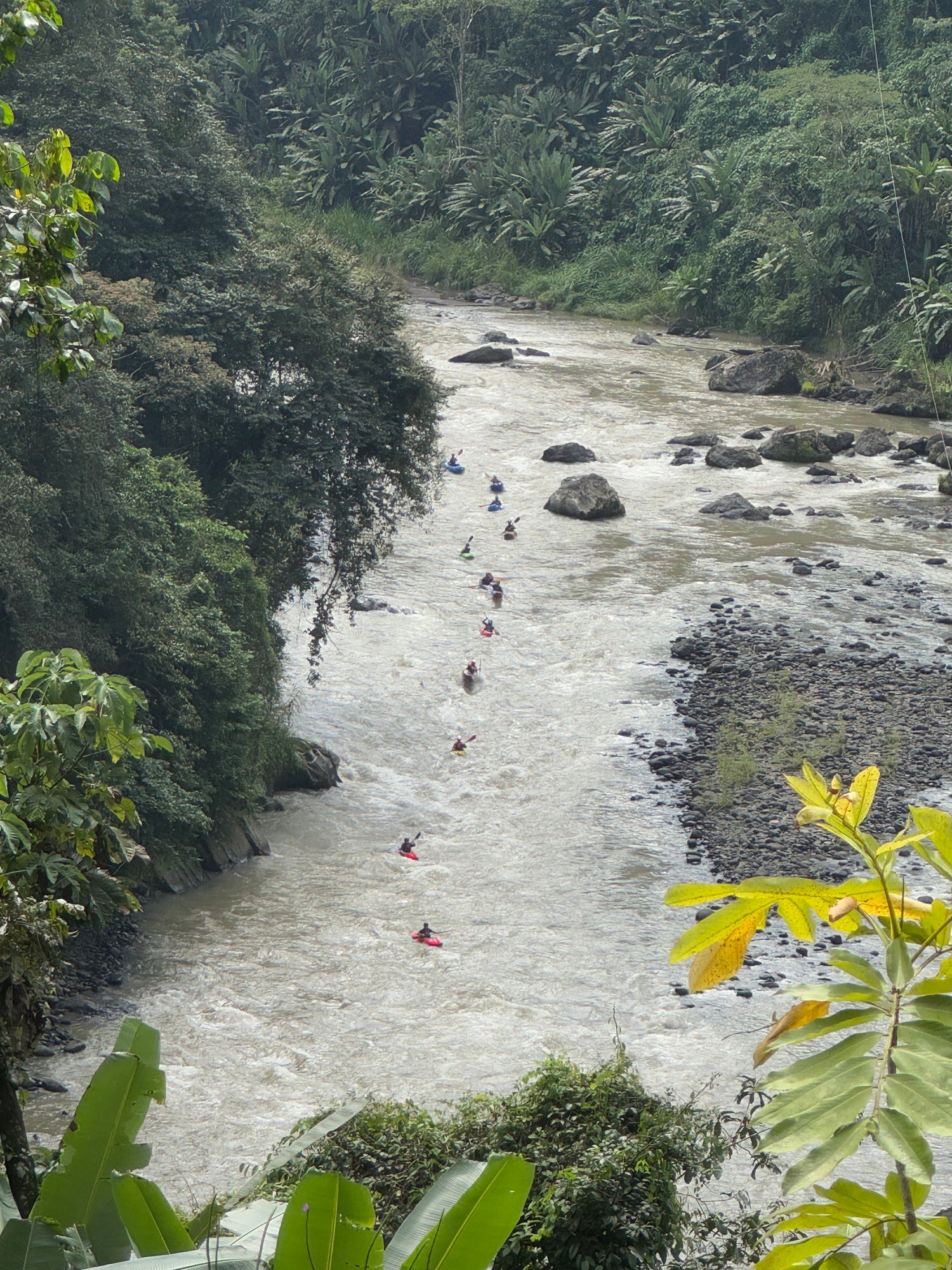 A group of kayakers enjoys an exhilarating Ladies Costa Rica Paddling Retreat by 4CRS Paddle School, paddling down a winding river amid lush greenery and rocks—a perfect adventure for women travel enthusiasts.