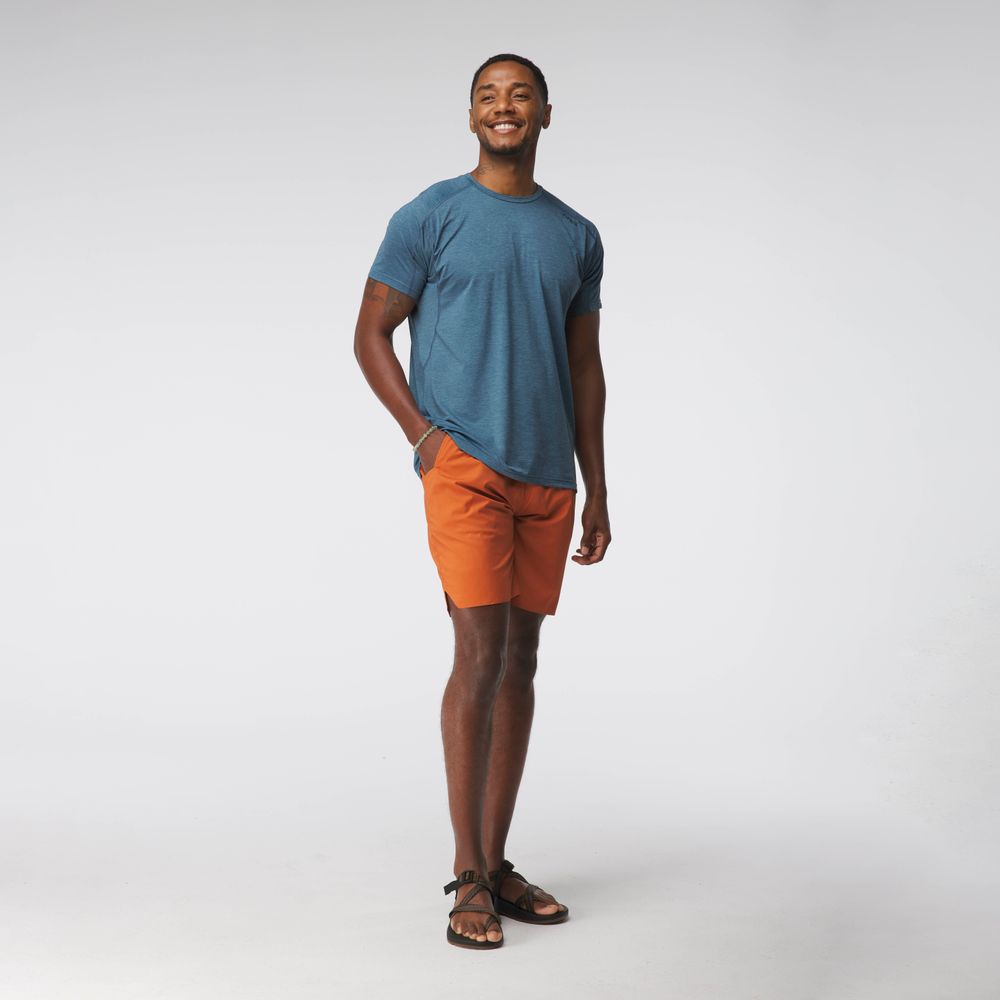 A man in an NRS Silkweight Short-Sleeve blue shirt and orange shorts stands smiling with hands in pockets against a plain background.