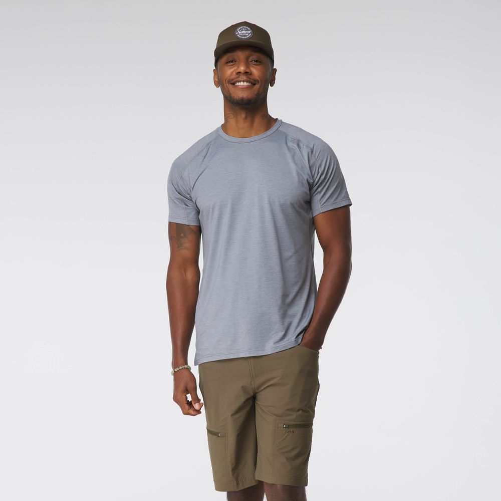 A man wearing an NRS Silkweight Short-Sleeve Shirt, khaki shorts, and a brown cap smiles against a plain background, ready for any adventure in his moisture-wicking attire.