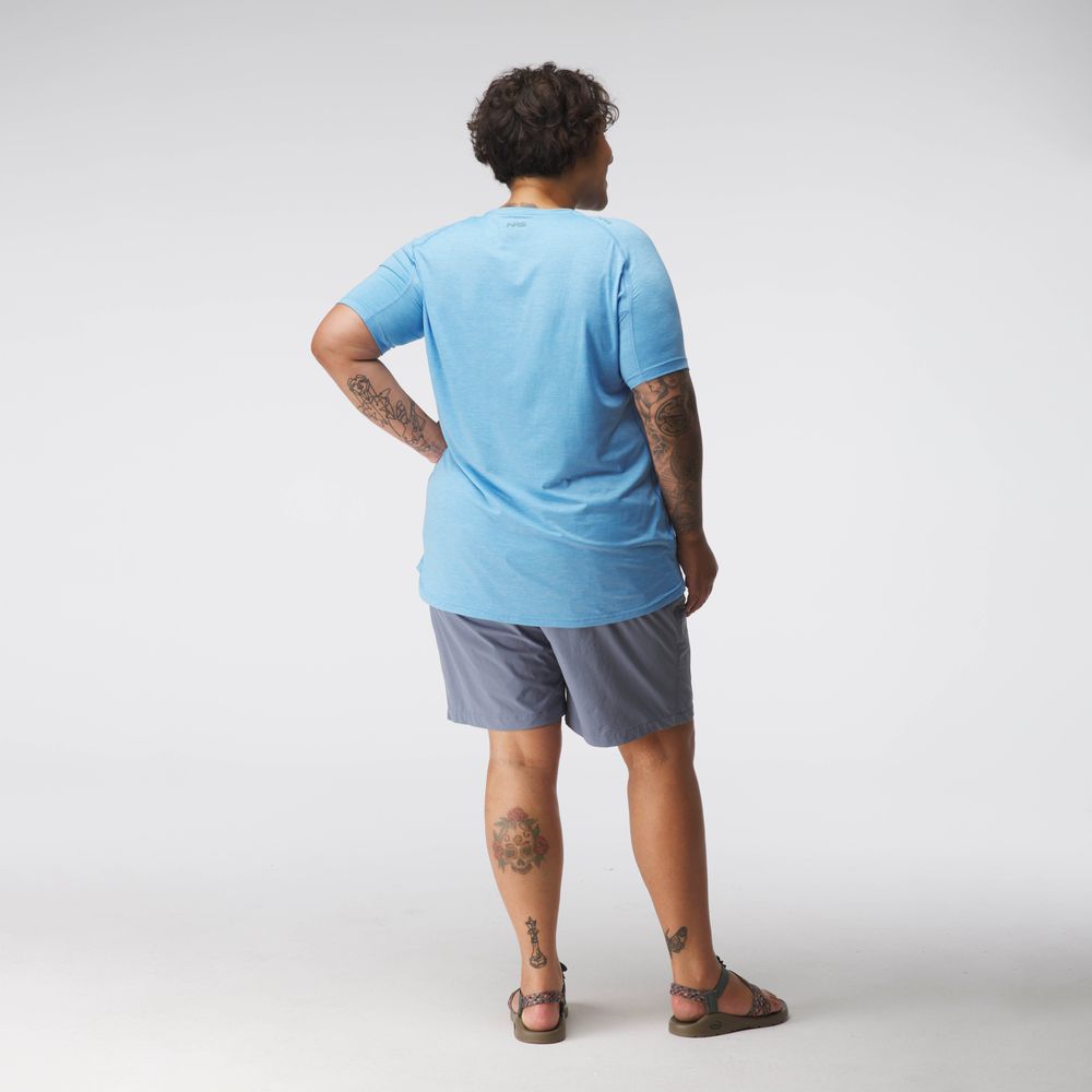 A person in an NRS Silkweight Short Sleeve Shirt in light blue and gray shorts stands back to the camera, revealing tattoos on arms and legs. The outfit ensures breathability against a neutral backdrop.