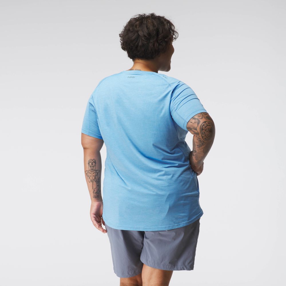 A person in a blue Silkweight Short Sleeve Shirt from NRS and gray shorts, seen from the back, stands confidently with tattooed arms and one hand on their hip. The outfit ensures breathability against the plain white backdrop.