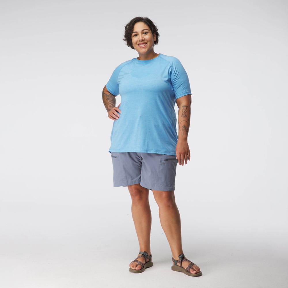 A person in a blue NRS Silkweight Short Sleeve Shirt, gray shorts, and sandals stands smiling against a plain background.