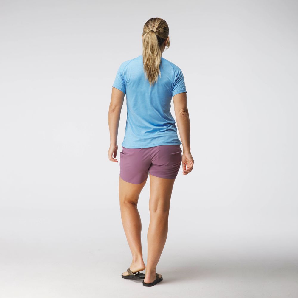 A person faces away from the camera wearing an NRS Silkweight Short Sleeve Shirt in blue with moisture-wicking fabric, purple sun-protective shorts, and black sandals against a plain background.