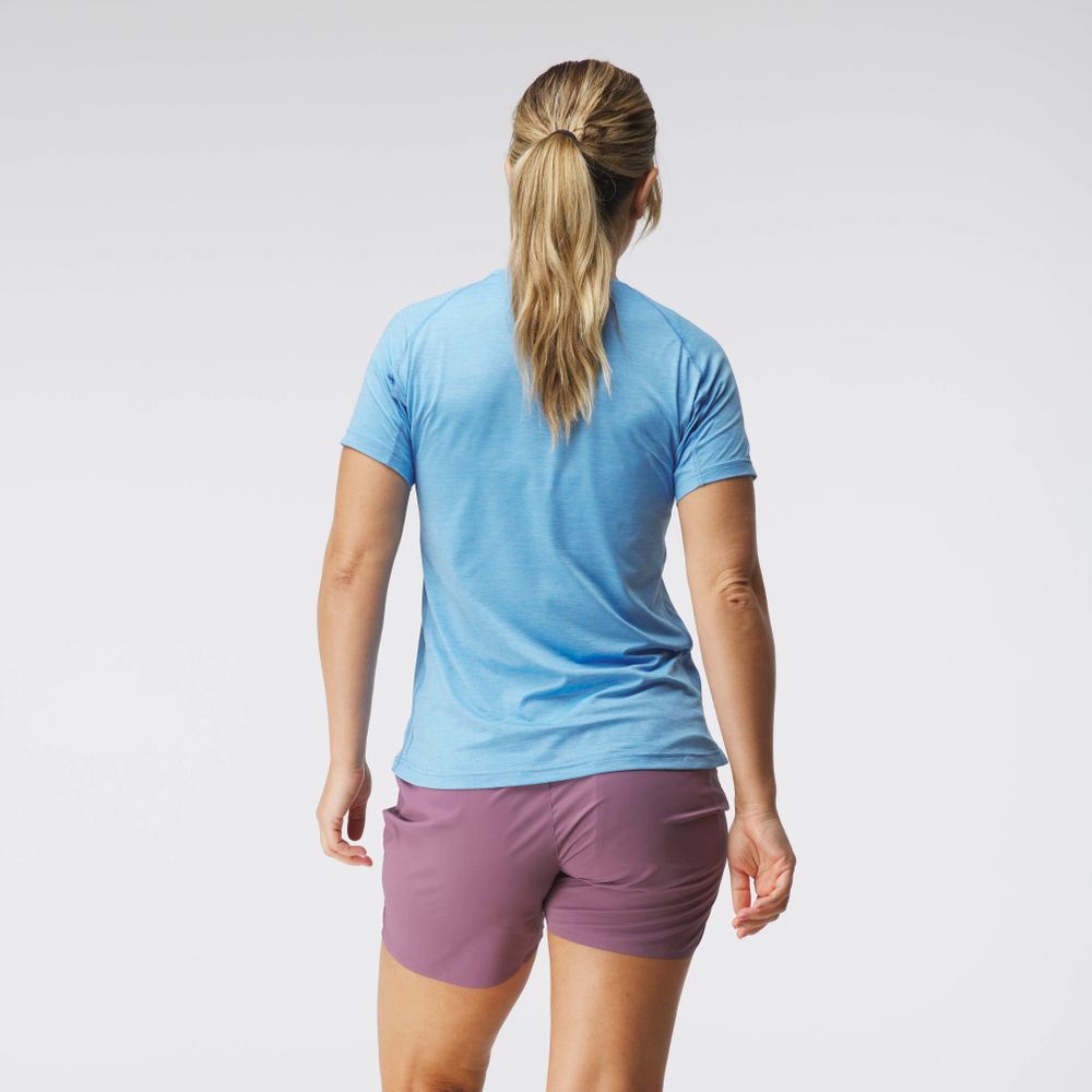 A woman with long hair tied in a ponytail, wearing the NRS Silkweight Short Sleeve Shirt in blue and purple shorts, stands facing away against a plain background.