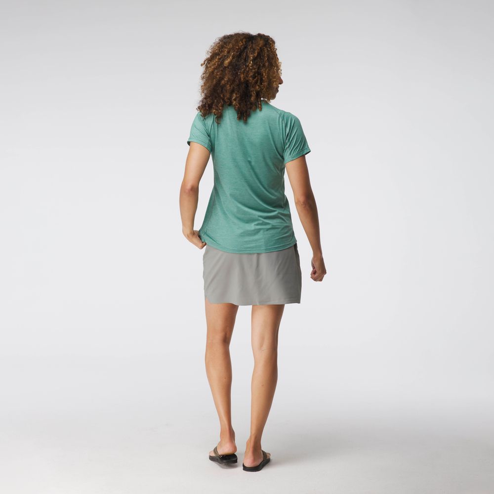 A person with curly hair is standing and facing away in a plain studio setting, wearing the NRS Silkweight Short Sleeve Shirt - Women's known for its breathability, paired with a gray skirt and flip-flops.