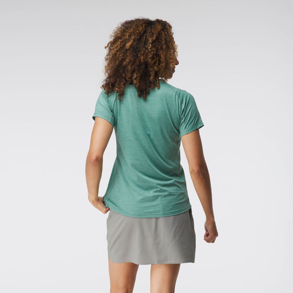 A curly-haired person wearing the breathable, moisture-wicking NRS Silkweight Short Sleeve Shirt in green and a gray skirt faces away against a plain background.