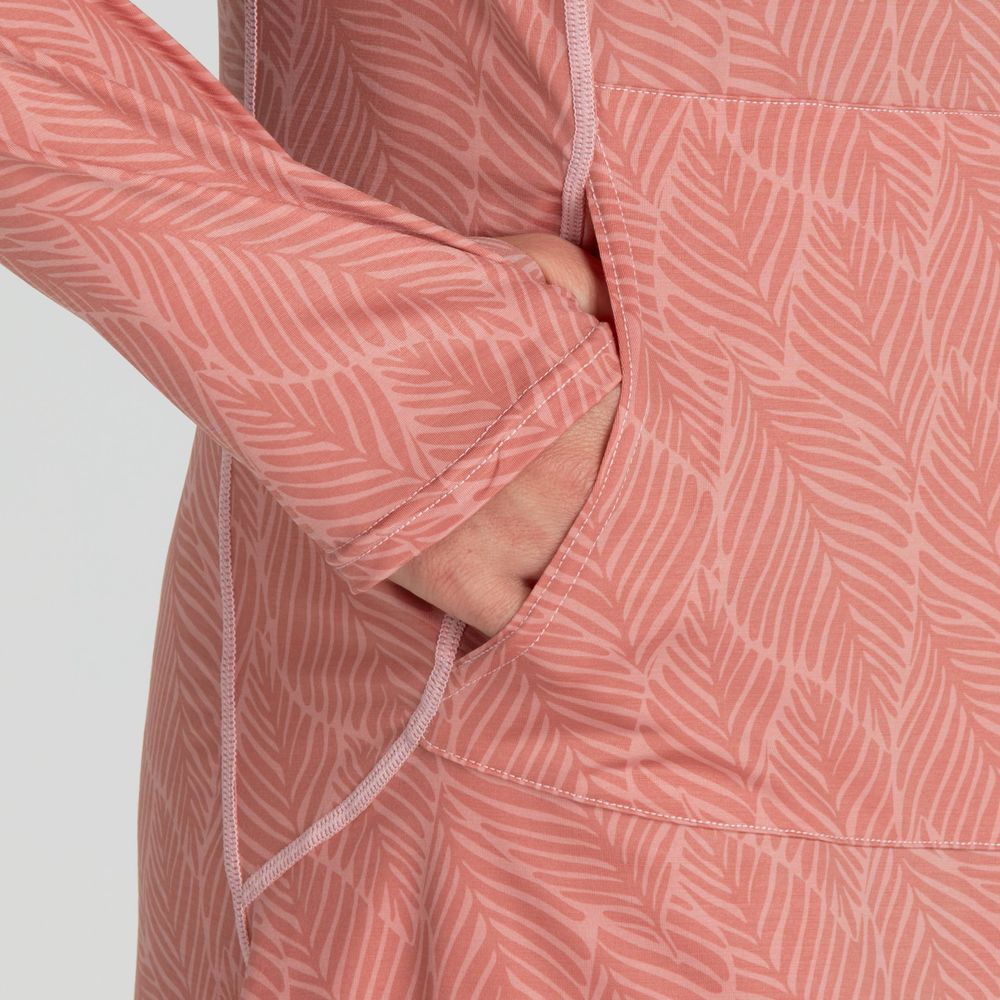 A close-up showcases a person wearing the NRS Silkweight Hoodie Dress—a long-sleeve pink top featuring a leaf pattern, crafted from recycled material. The hand stylishly tucked in a side pocket adds both flair and practicality.