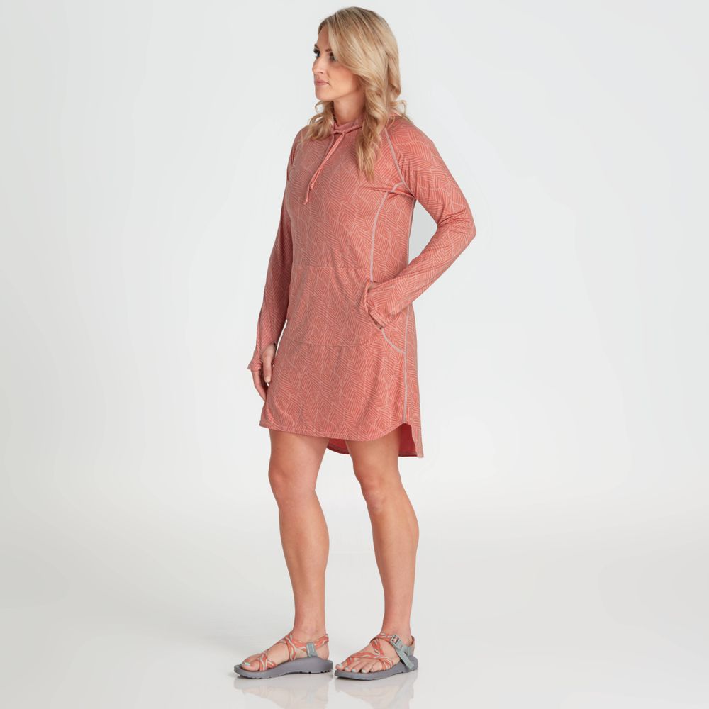 A person stands sideways in sandals, wearing the salmon-colored NRS Silkweight Hoodie Dress, a long-sleeve hooded dress with a geometric pattern made from recycled material, effortlessly combining style with sun protection.