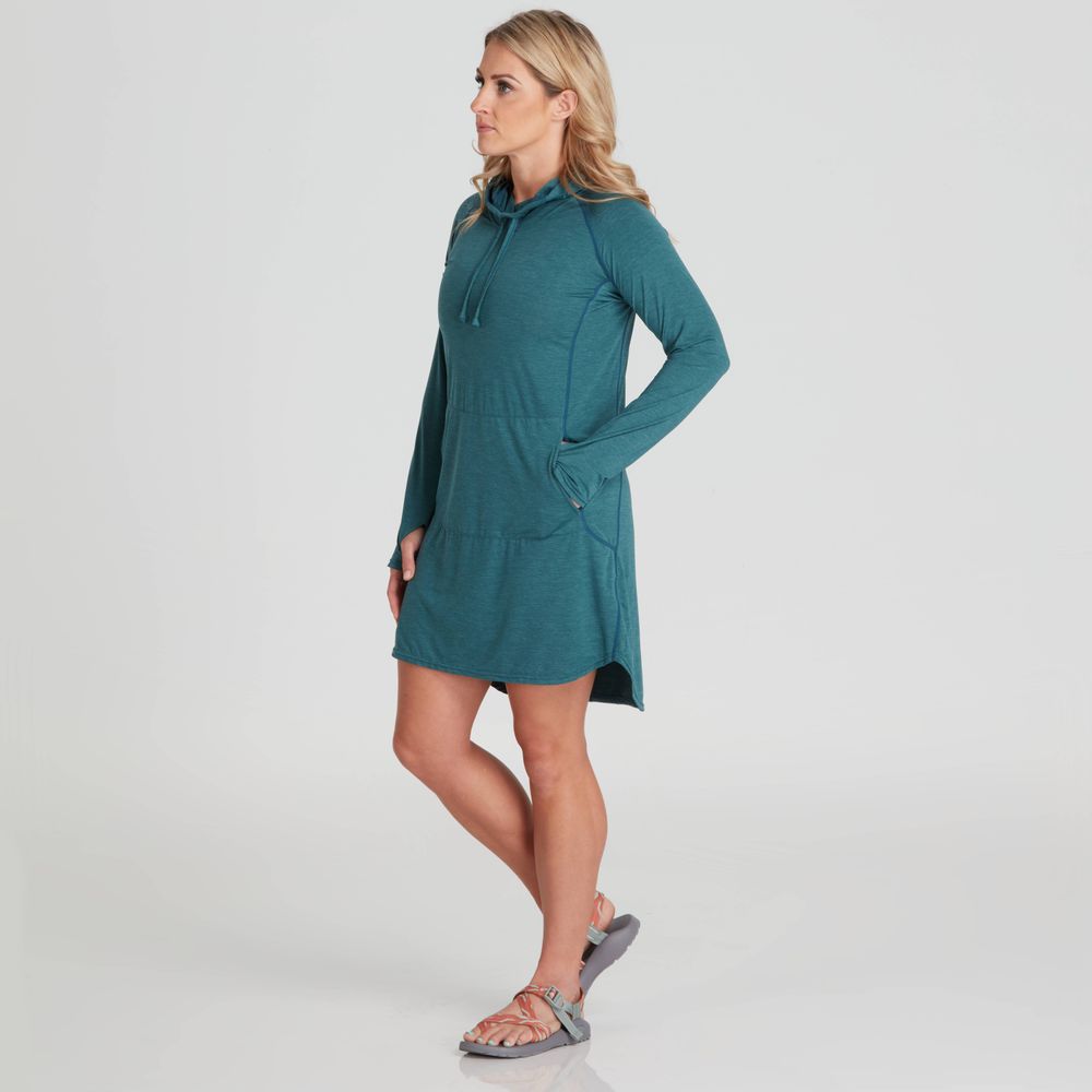 Wearing a teal NRS Silkweight Hoodie Dress with sandals, a person stands against a plain backdrop, looking sideways with hands in pockets, appreciating the sun protection from its recycled fabric.