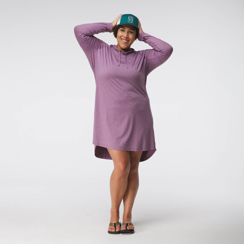 Wearing an NRS Silkweight Hoodie Dress in purple and sandals, a person smiles while holding a recycled green hat that provides excellent sun protection against a plain background.