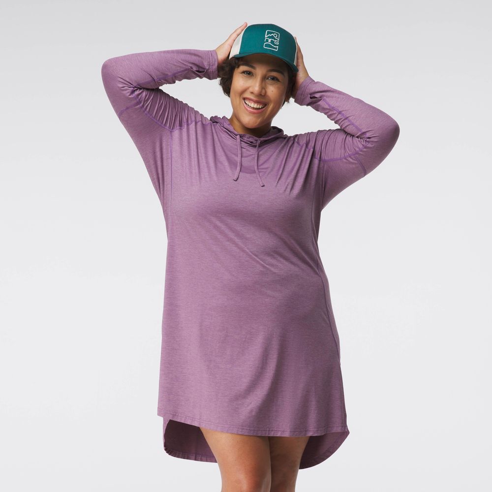 A person smiles, wearing an NRS Silkweight Hoodie Dress crafted from recycled materials and a dark cap, adjusting the hat to shield from the sun.
