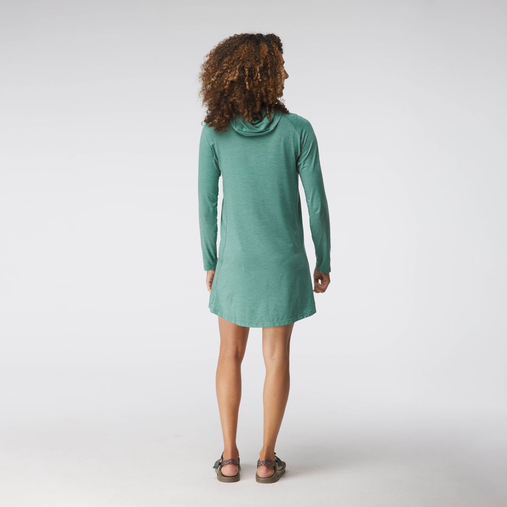 A person with curly hair elegantly wears the NRS Silkweight Hoodie Dress, a green long-sleeve piece made from recycled materials. Facing away from the camera on a plain background, this dress blends elegance and sustainability.