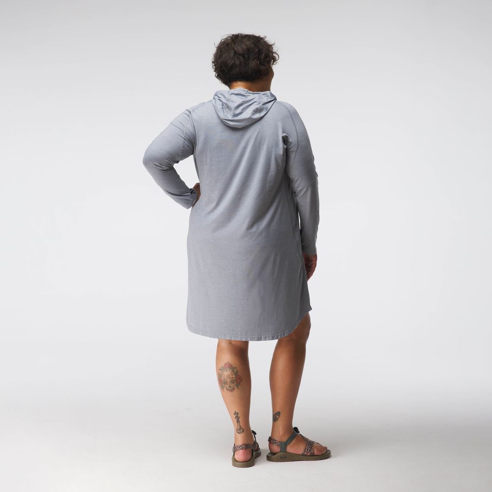 A person with their back to the camera wears a light gray NRS Silkweight Hoodie Dress made from recycled materials, paired effortlessly with sandals. Tattoos peek out on their legs, adding artistic flair.