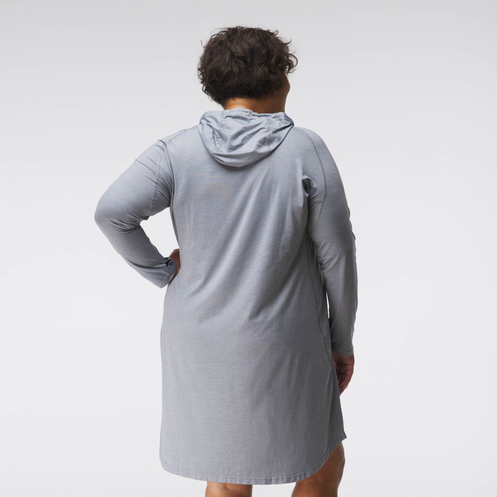 A person in an NRS Silkweight Hoodie Dress, crafted from recycled materials, stands with their back to the camera in soft light, adding style to the neutral space and offering subtle sun protection.