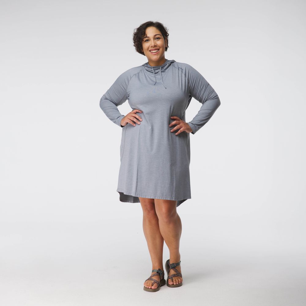 A smiling person with short curly hair, wearing NRS's Silkweight Hoodie Dress in gray made from recycled material and sandals, stands against a plain white background.