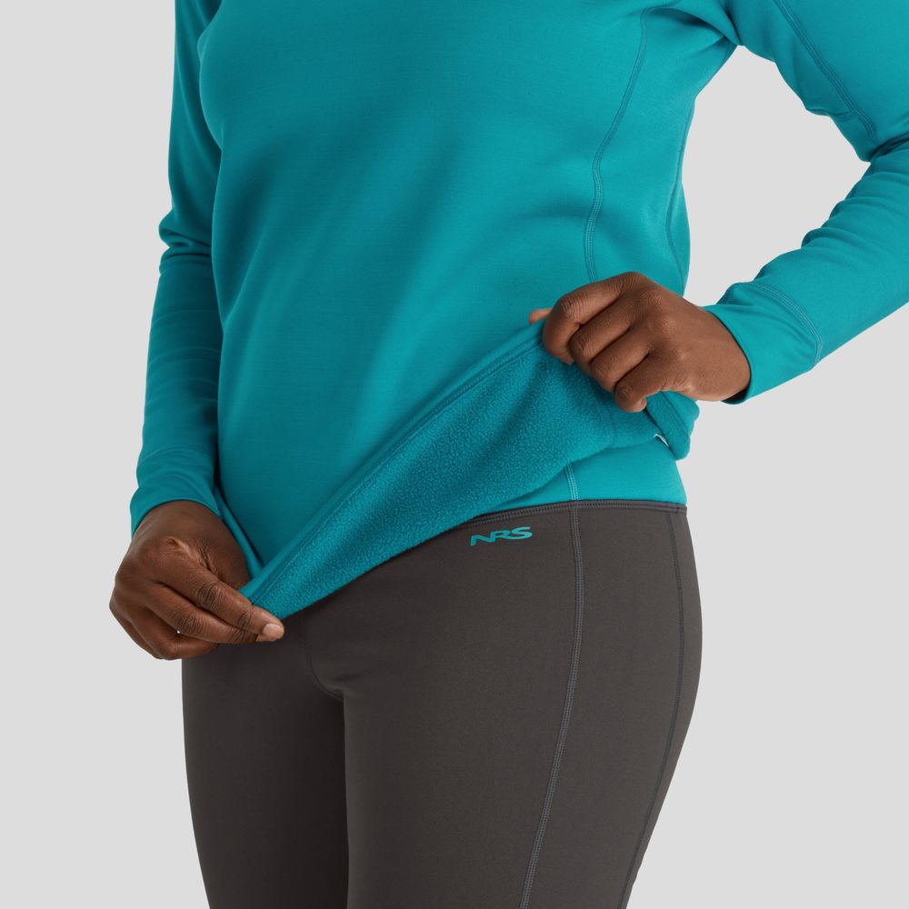 The person shows off their teal long-sleeve NRS Expedition Fleece top and gray leggings, highlighting its recycled inner lining. This women's top offers UPF 50+ protection, combining sustainability with outdoor-ready style.