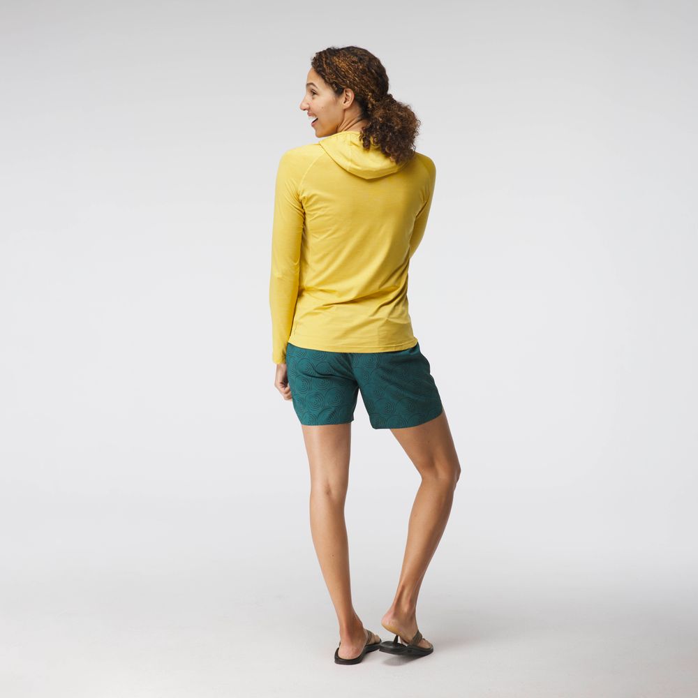 Wearing a NRS Silkweight Hoodie - Women's in vibrant yellow and green shorts, the person stands in a relaxed pose, looking over their shoulder against a plain background.