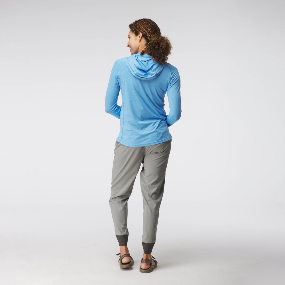 A person with curly hair stands against a light background, wearing gray pants, sandals, and a blue NRS Silkweight Hoodie - Women's, designed with technical fabrics for comfort and sun protection.