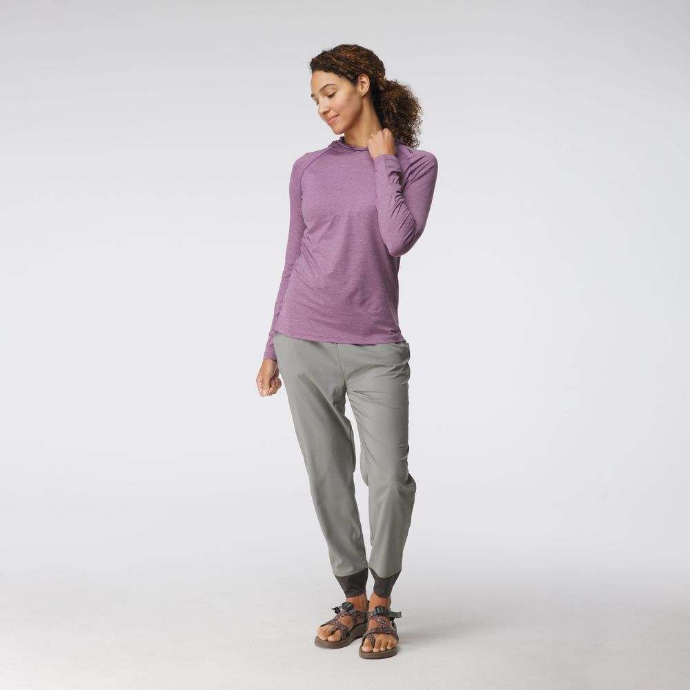 A woman in a purple NRS Silkweight Hoodie and gray pants stands against a plain background, looking down with a slight smile.