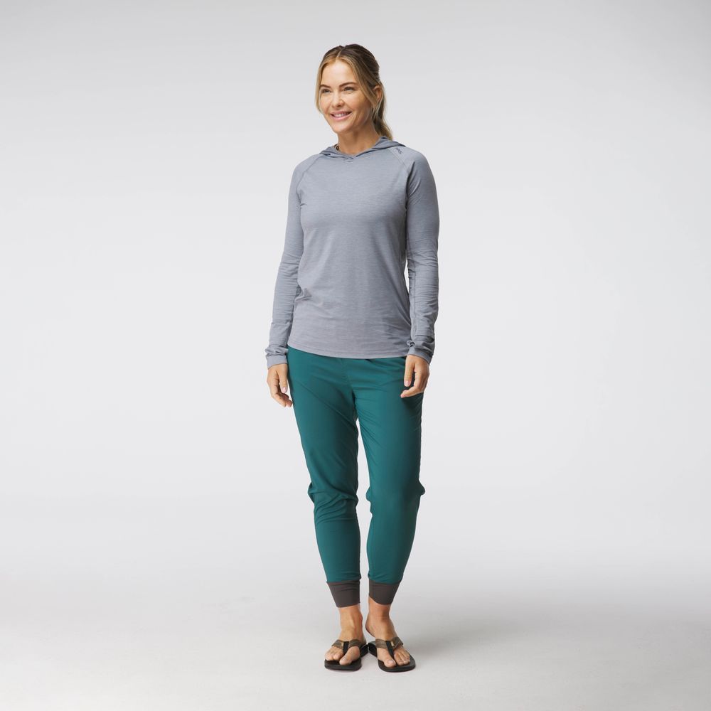 A woman in a casual gray NRS Silkweight Hoodie and teal pants smiles in a studio setting, wearing black sandals. Her light brown hair is pulled back, showcasing the comfort and sun protection of technical fabrics.