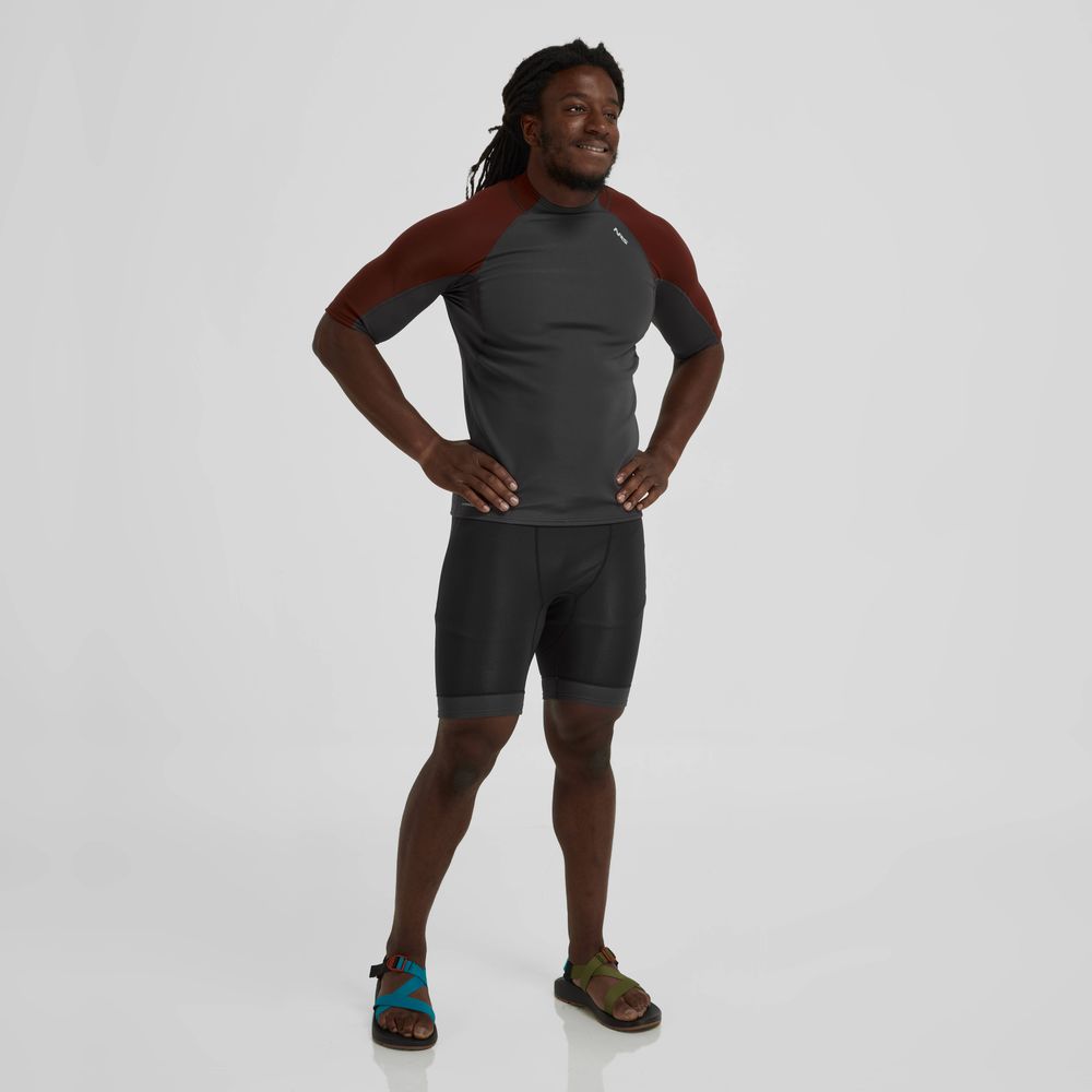 A man stands confidently in a dark NRS Hydroskin 0.5mm Shorts for men, paired with neoprene sandals, against a neutral background.