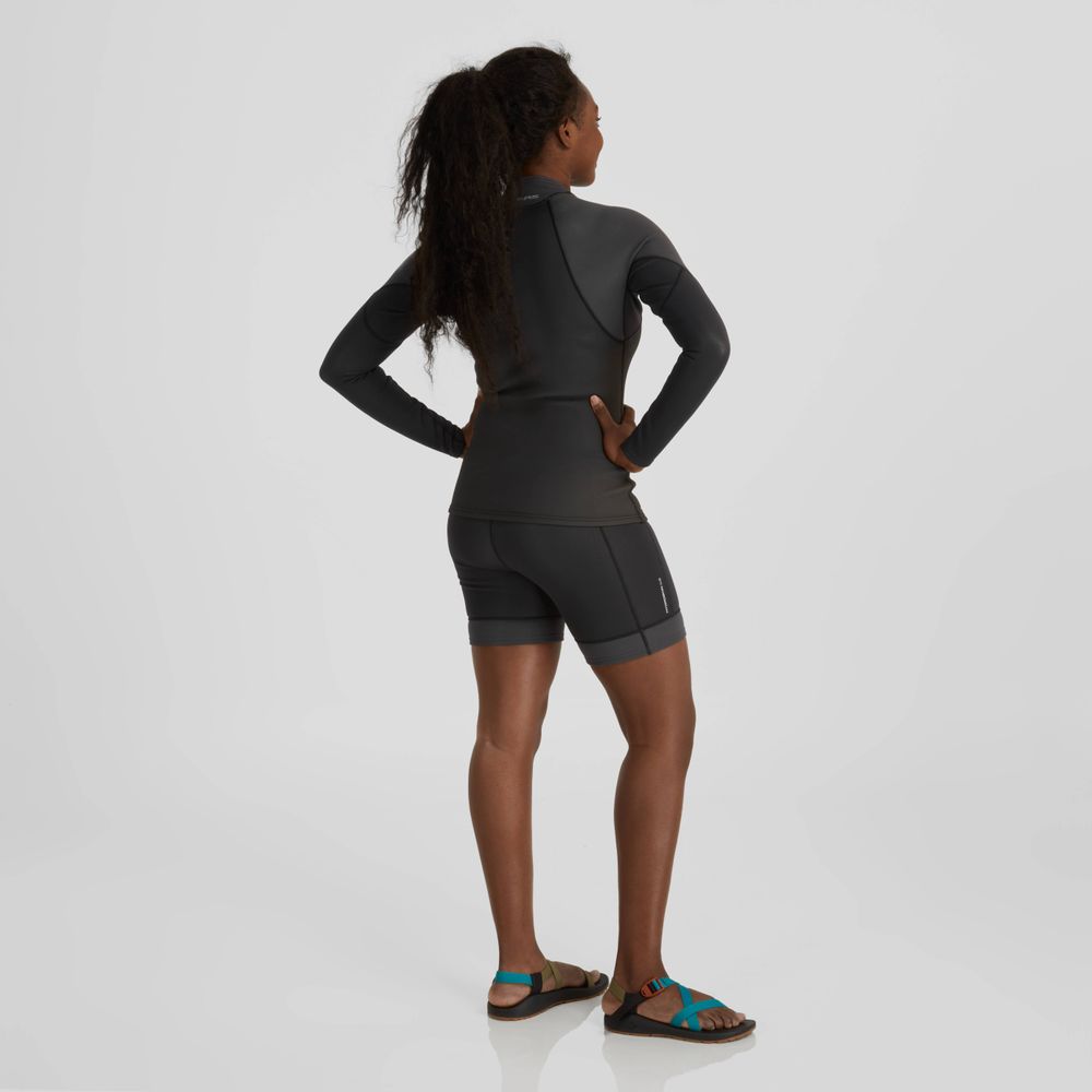 With their back turned, the person wears an NRS HydroSkin 0.5mm Shorts - Women's in sleek black Terraprene neoprene and comfortable sandals, poised for paddling performance on a plain background to enhance agility and comfort.