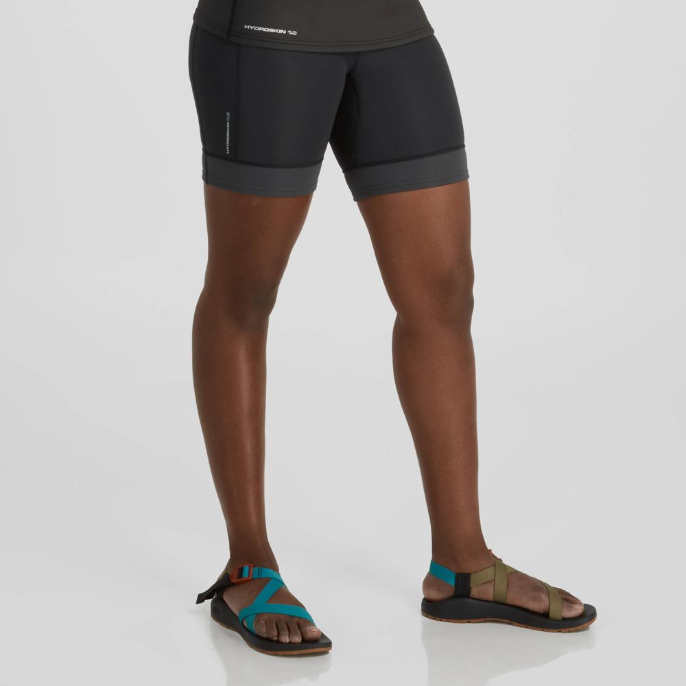 A person in NRS HydroSkin 0.5mm Shorts - Women's and colorful sandals stands against a plain backdrop, highlighting their commitment to paddling. Only the lower body is visible, showcasing Terraprene neoprene for flexibility and comfort.