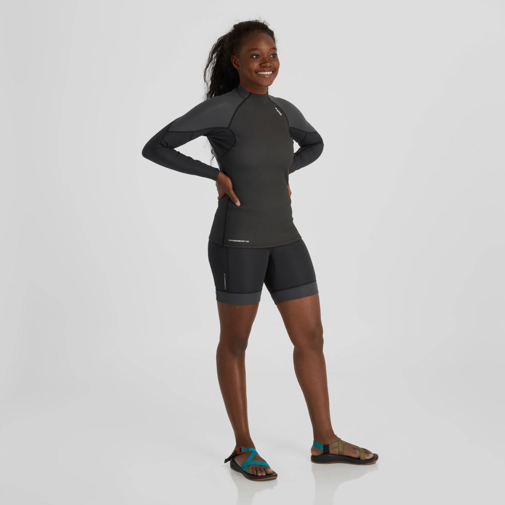 A person wearing a dark long-sleeve rash guard and NRS HydroSkin 0.5mm Shorts - Women's stands smiling in performance paddling sandals against a plain white background.