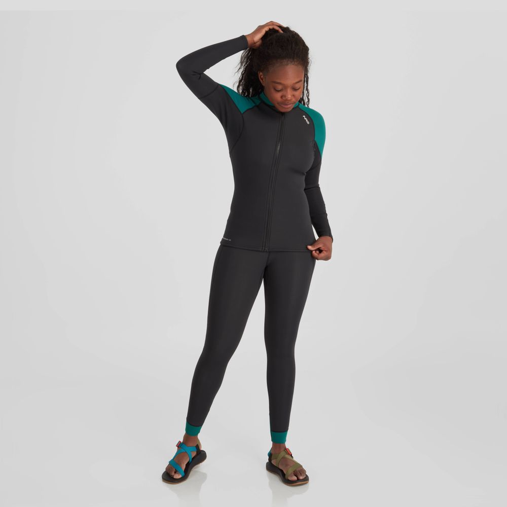 A woman, dressed in NRS Hydroskin 1.5mm Pants with a black and teal pattern and neoprene insulation, is paired with sandals. She stands on a plain background, looking down thoughtfully with one hand on her head, ready for optimal paddling performance.