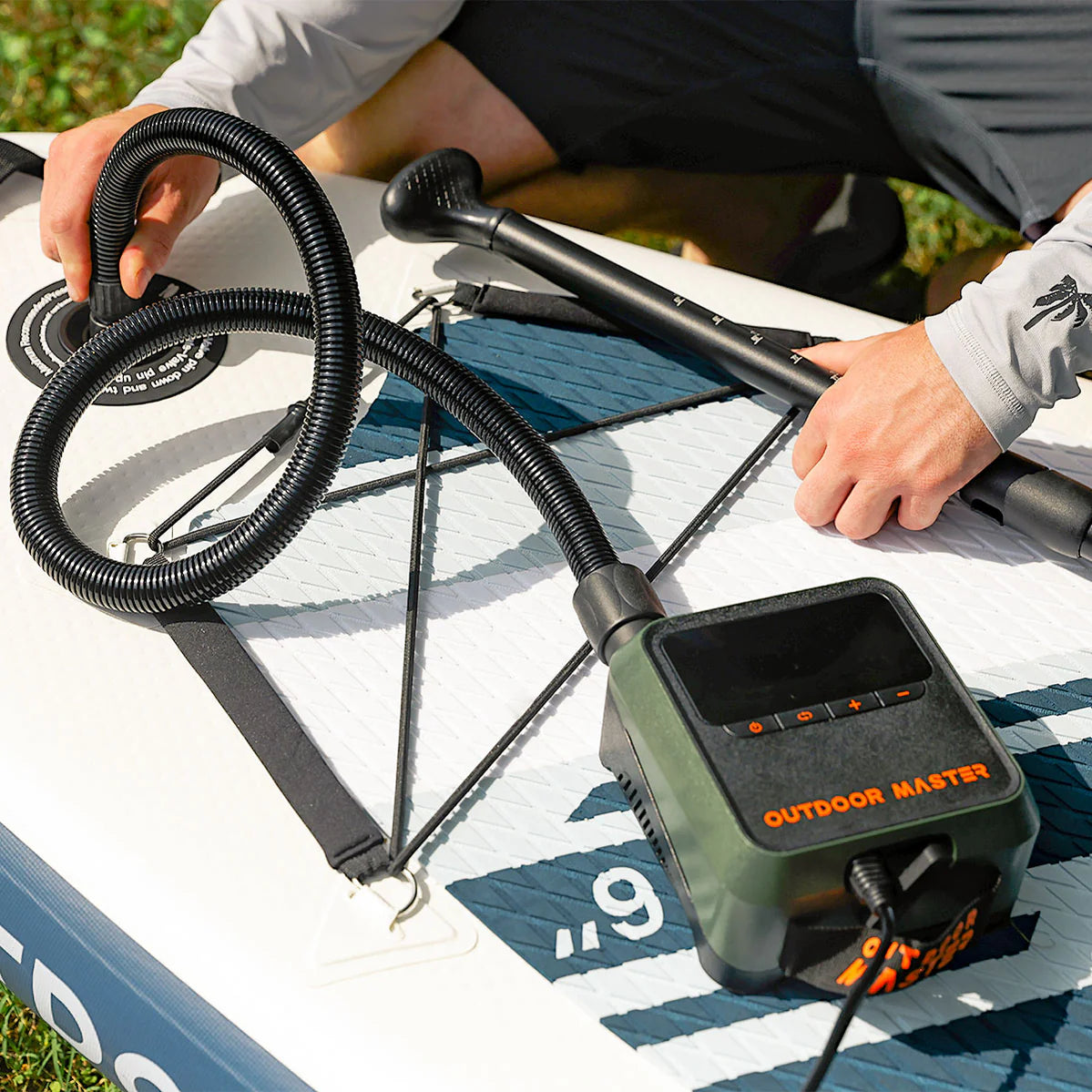 A person using the Outdoor Master Cachalot 2 - 12V Mini Electric SUP Pump to inflate a stand-up paddleboard outdoors.