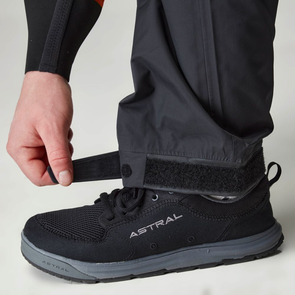 Someone adjusts the Velcro strap on a black shoe, wearing NRS Endurance Splash Pant for men, crafted from breathable HyproTex fabric to ensure dryness and comfort.