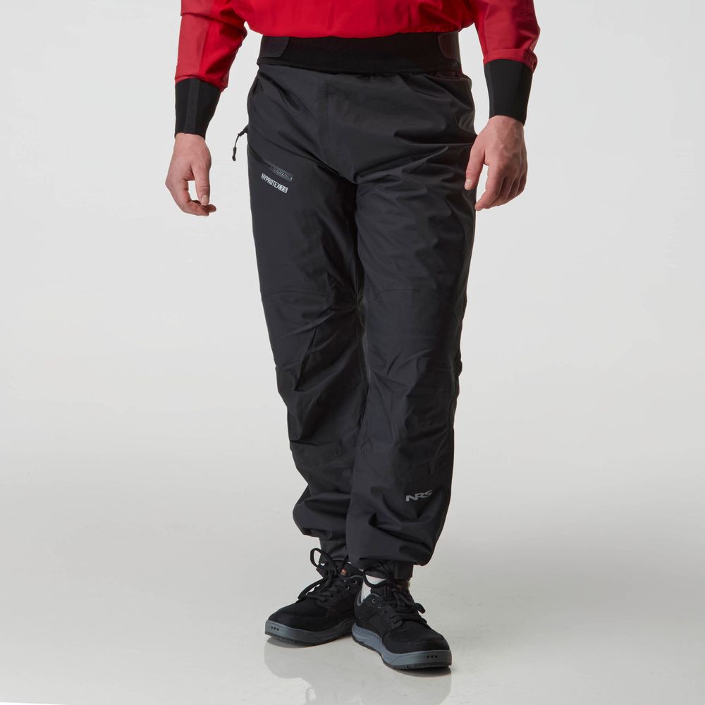 A person stands against a plain background wearing NRS's Endurance Splash Pant for men, which features a black design with reinforced seat and knees, paired with a red top and matching black shoes.