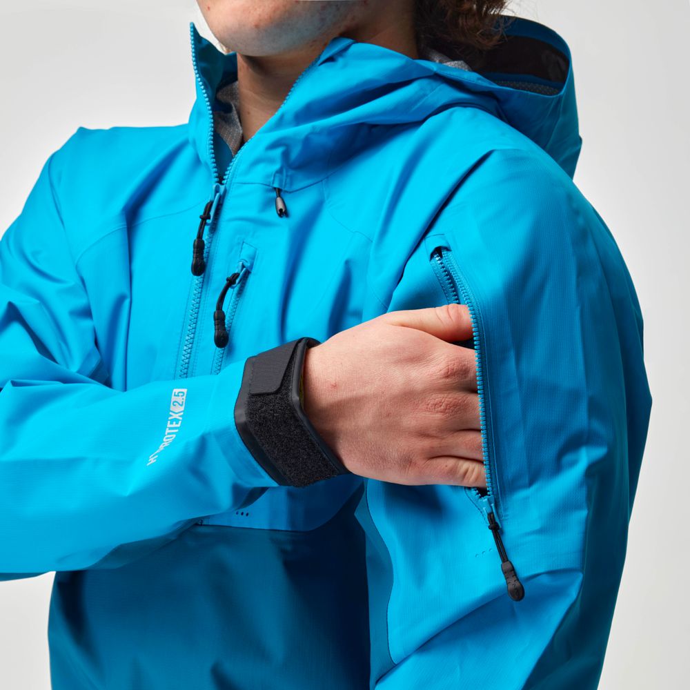 A person wearing the NRS High Tide Jacket in bright blue adjusts the zipper on a sleeve pocket. This waterproof splash jacket features an adjustable hood for optimal paddling protection.