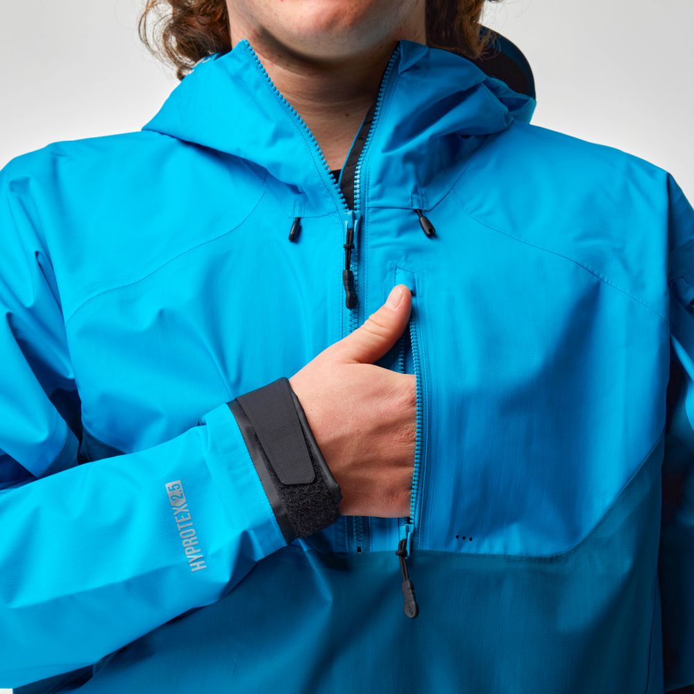 A person wearing the NRS High Tide Jacket in bright blue adjusts the zipper, while its waterproof breathable design and adjustable hood offer extra protection.