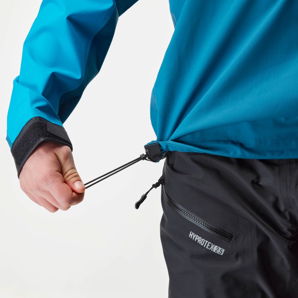 A person adjusts the drawstring on an NRS High Tide waterproof blue splash jacket, paired with dark pants featuring a zip pocket labeled "HYDROTEK 2.5," ideal for paddling protection with its adjustable hood.