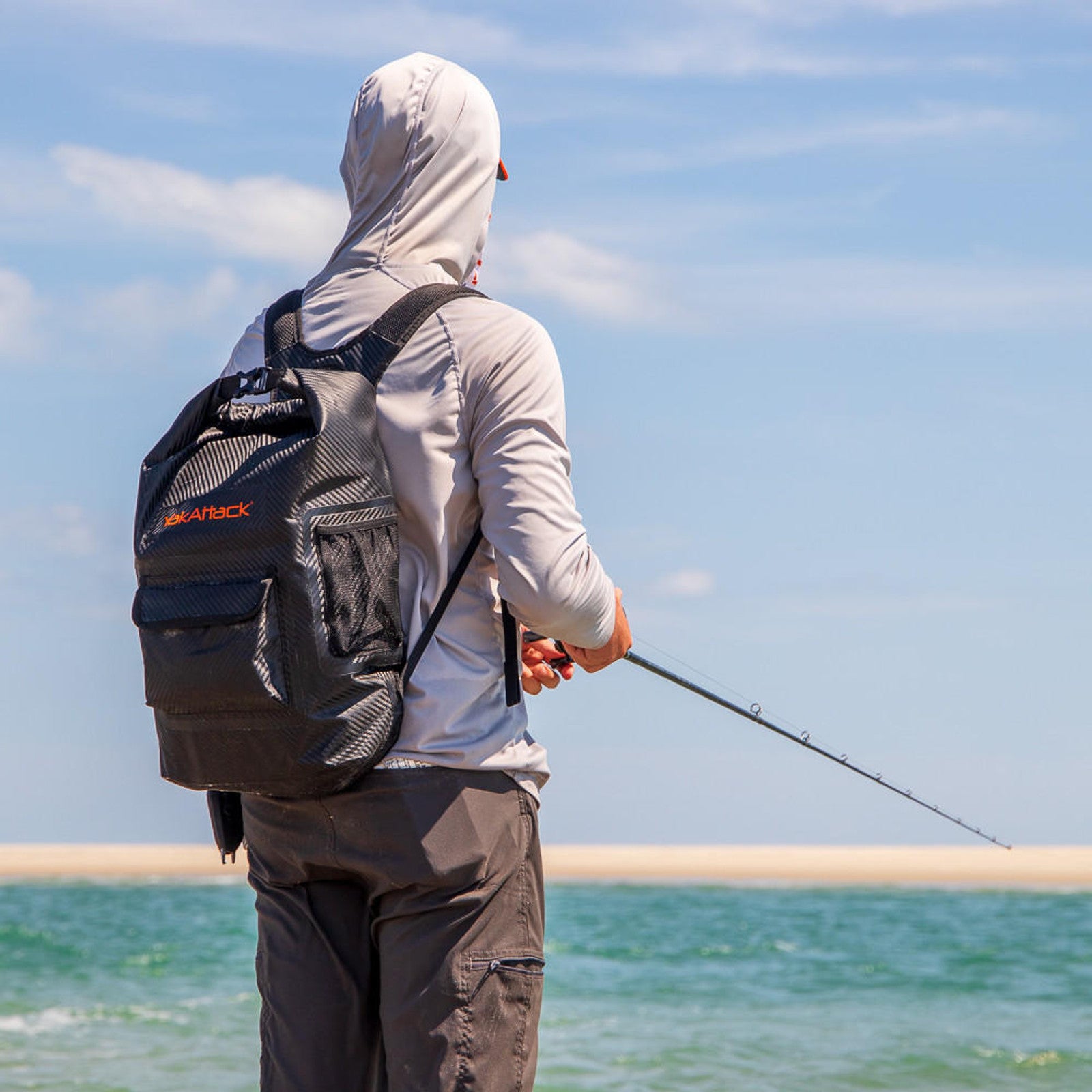 A person with a YakAttack 20L Roll-Top Drybag Backpack and a gray hooded shirt stands on a sandy beach, fishing with a rod while facing the water under a blue sky.