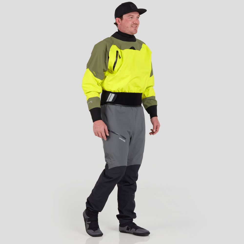 A person stands smiling against a plain backdrop, showcasing paddling performance in an NRS REV (GORE-TEX Pro) drytop, bright yellow and gray, paired with black boots and cap.