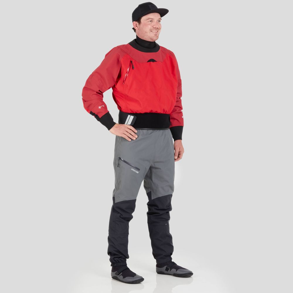An individual wearing the NRS REV (GORE-TEX Pro) Drytop in red and gray stands against a light gray background, prepared for peak paddling performance.