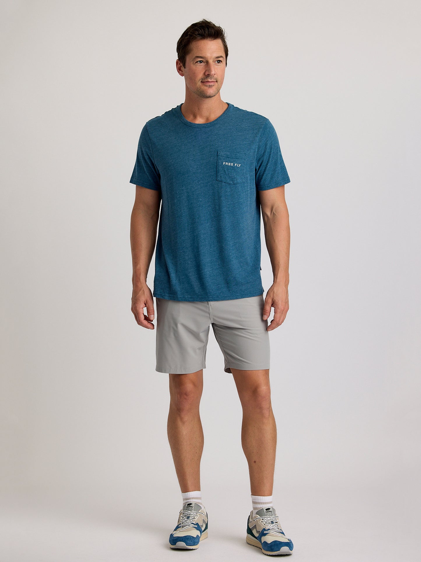 A man faces forward wearing the Free Fly Salt and Sun Pocket Tee M's by Free Fly, paired with light gray shorts and blue and white athletic shoes, set against a plain light background.