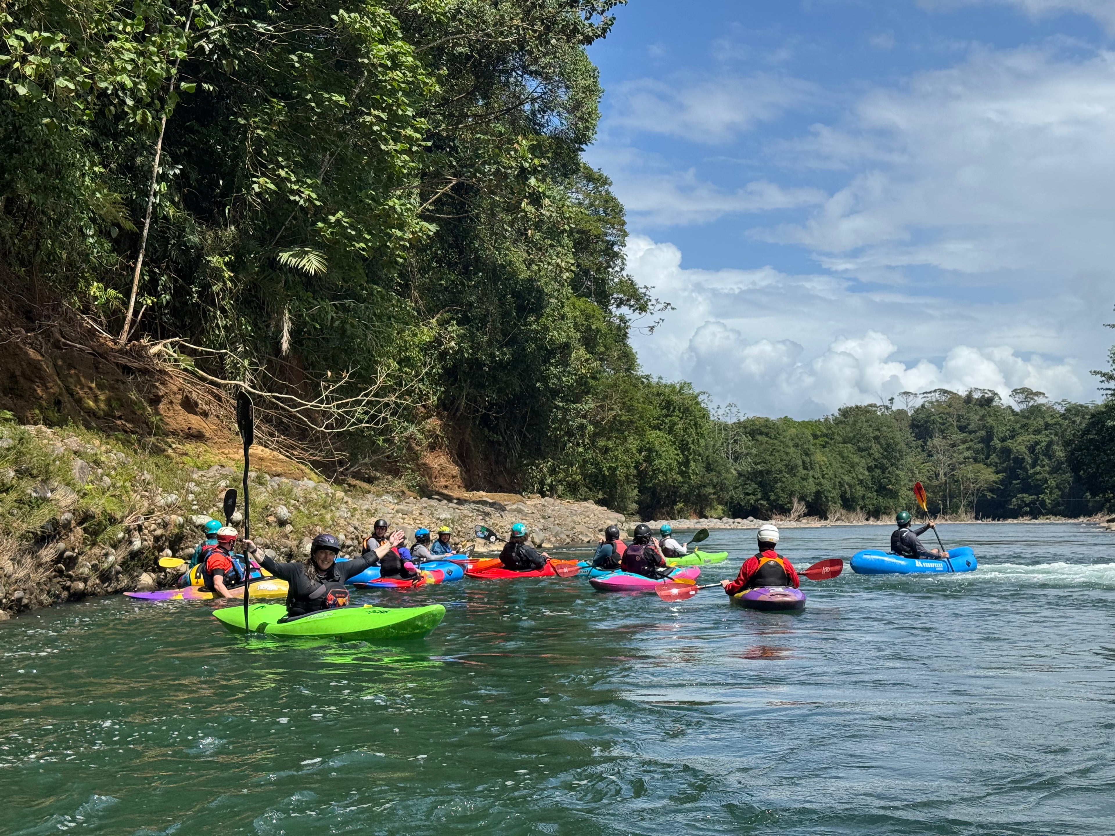 A group of women enjoy paddling colorful kayaks on a scenic river surrounded by lush trees under partly cloudy skies during the 4CRS Paddle School Ladies Costa Rica Paddling Retreat.
