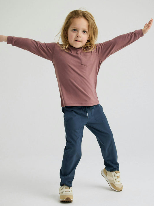A child with long hair stands with arms outstretched, wearing a Free Fly Toddler Bamboo Shade Hoody offering UPF 50+ sun protection, along with dark pants and light-colored shoes against a plain background.