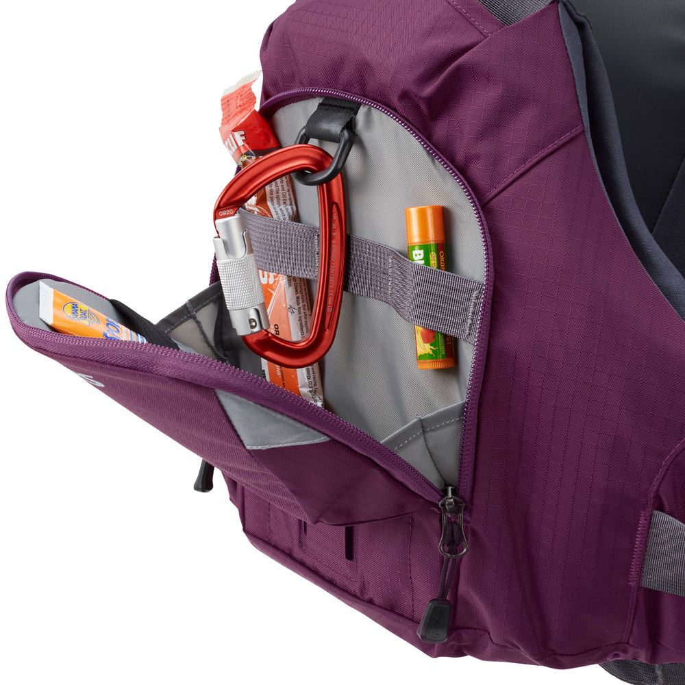 A purple backpack with an open front pocket resembles the NRS Ninja PFD, containing a red carabiner, green lip balm, and a snack bar. The pocket is equipped with a gray strap for organization.