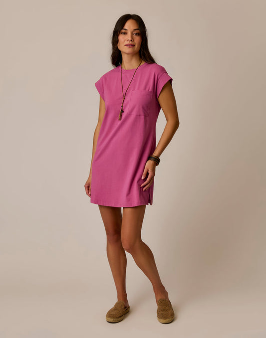 A woman stands against a plain background wearing the Carve Cosmo Dress, a vintage-inspired, short-sleeve pink organic cotton piece, styled with a long necklace, bracelets, and beige slip-on shoes.