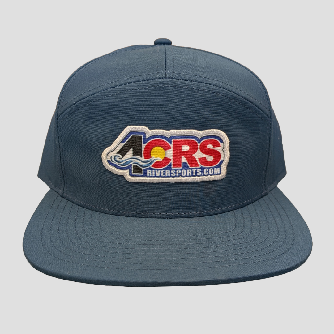 The 4CRS 7 Panel Performance Hat by 4Corners Riversports is a blue baseball cap with the "4CRS RIVERSPORTS.COM" logo on the front and a wave design in blue, red, and yellow.