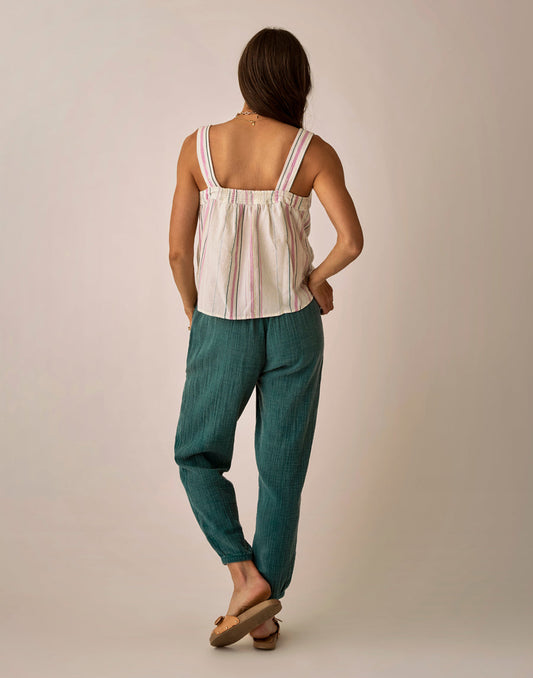 Woman facing away from the camera, wearing Carve's Calista Gauze Pant in teal, a sleeveless striped top, and tan sandals against a plain background.