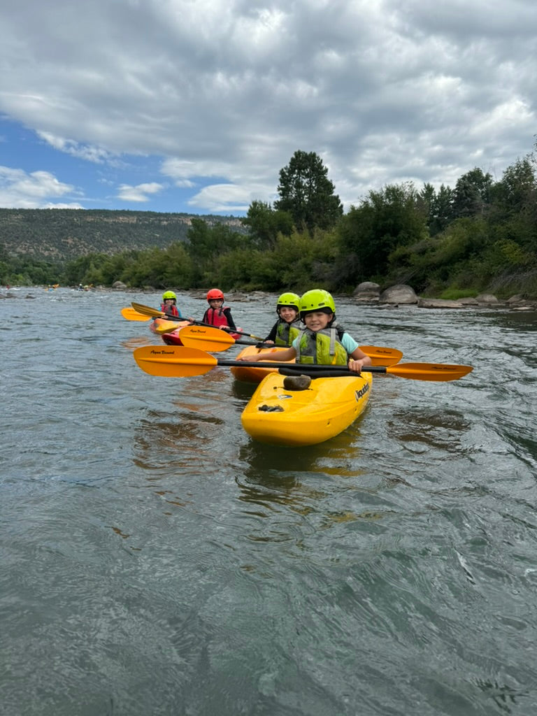 Four kids in helmets and life jackets paddle yellow kayaks on a scenic river—a fun adventure with 4CRS’s Kids Paddle School Class Holiday Gift Certificate. Perfect for young paddlers who love the outdoors!.
