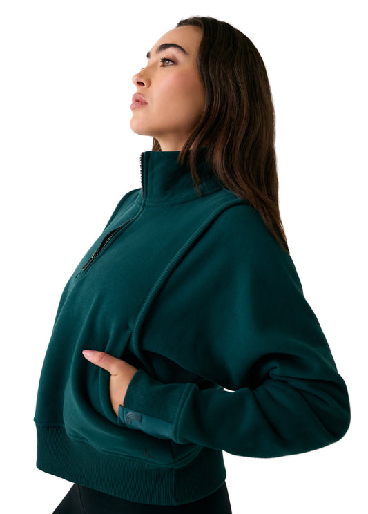 A woman stands in profile with her left hand in the pocket, wearing the Lole Yonder Half Zip cropped sweatshirt in dark green, set against a white background.