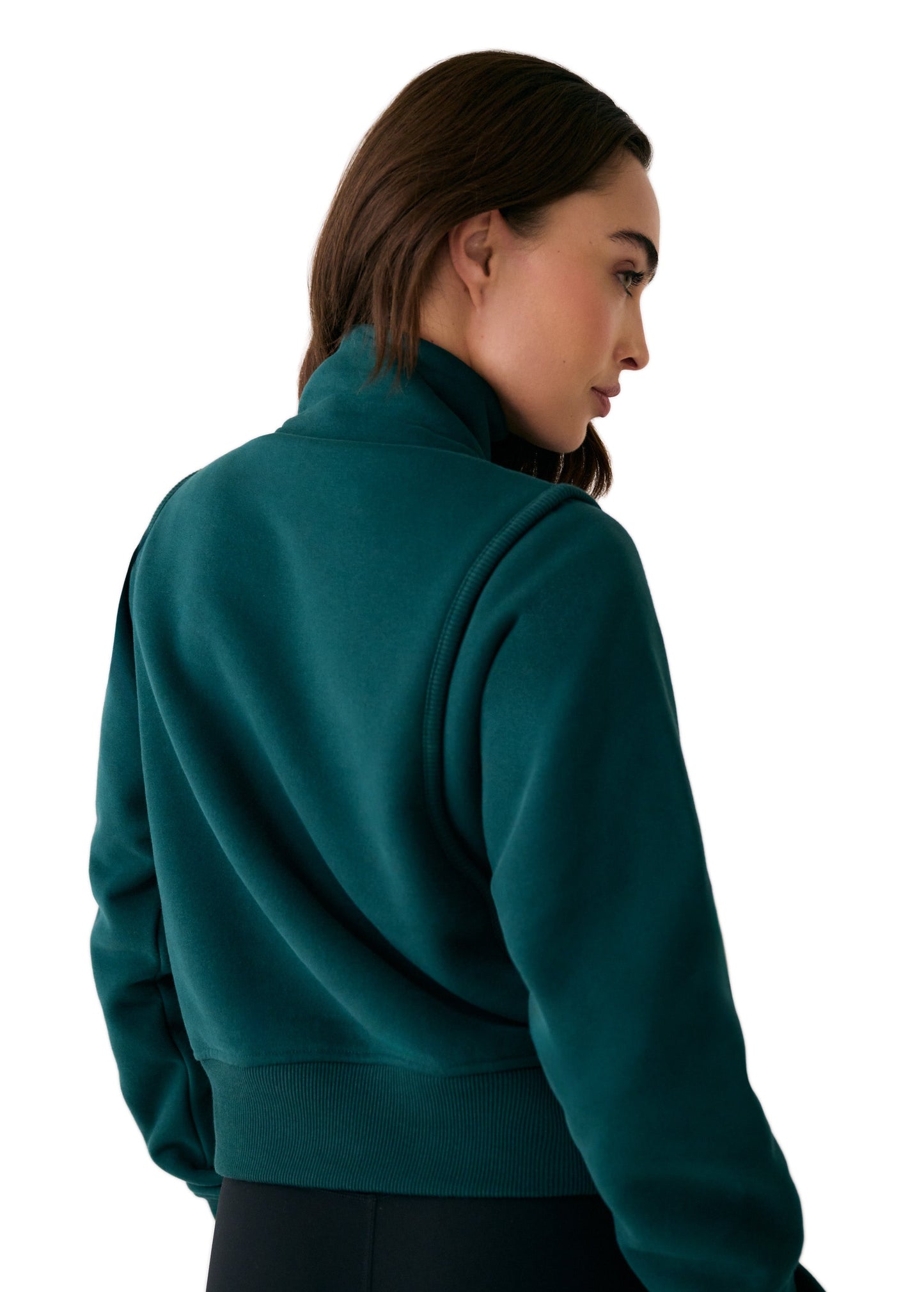 A woman with long brown hair is shown from the back, wearing the Lole Yonder Half Zip in dark green and black pants, standing against a white background.