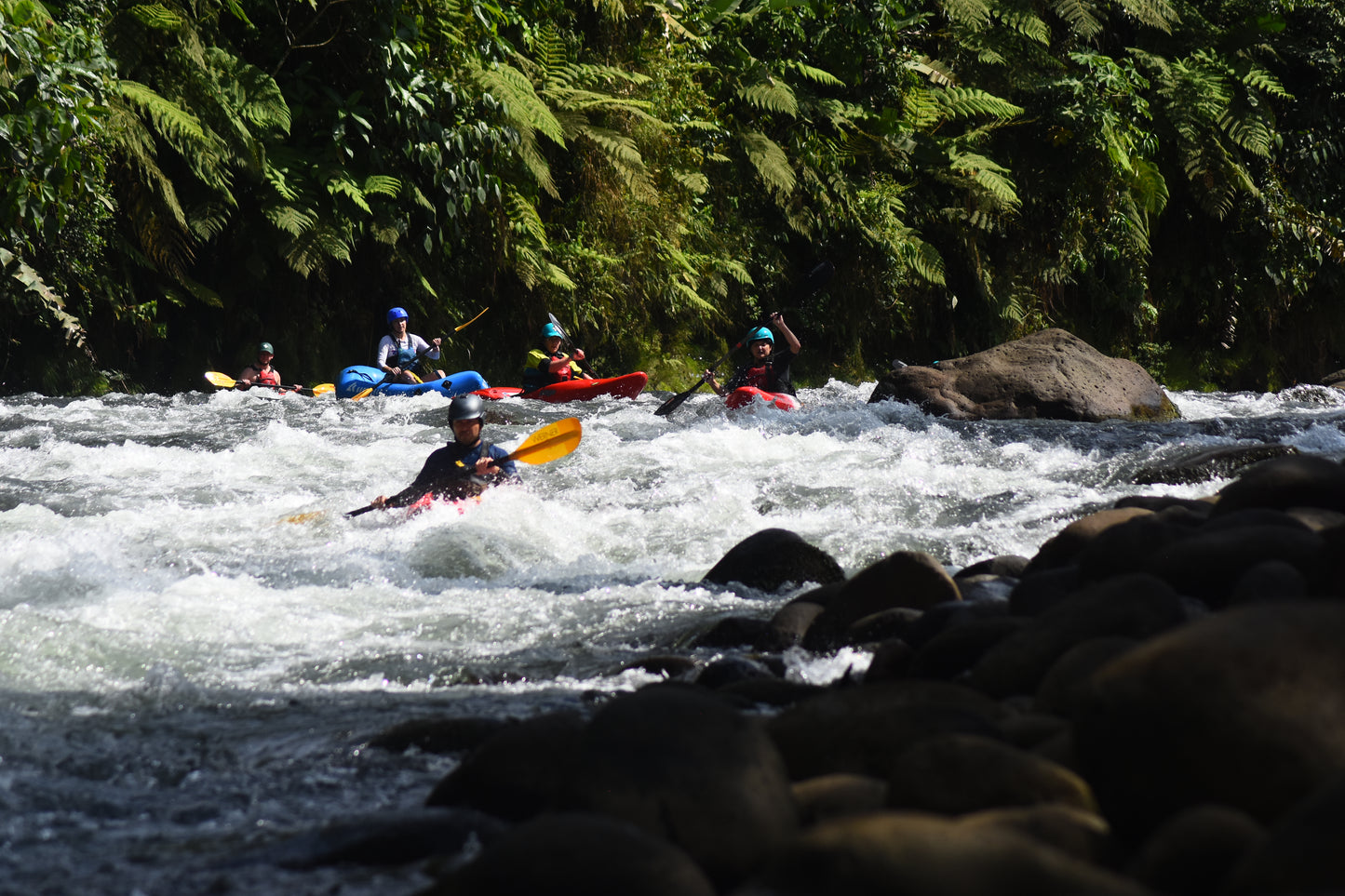 Five kayakers from 4CRS Paddle School navigate through rapids in a river surrounded by lush green vegetation and rocks during the Costa Rica Paddling Trip.