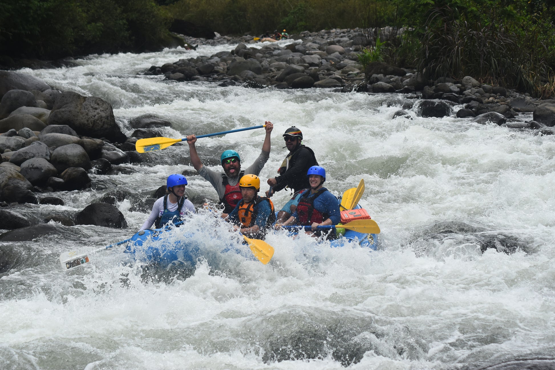 A group of people wearing helmets and life jackets are white-water rafting down a rocky river with one person raising a paddle in the air, showcasing their skills learned during the Costa Rica Paddling Trip offered by 4CRS Paddle School.